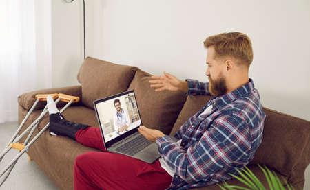 Man with leg injury while sitting at home talking to doctor on video call during online...