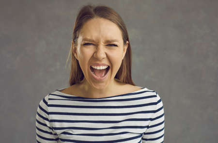 Close up studio shot of a crazy stressed beautiful young woman with an angry displeased face expression screaming loudly at the camera. Portrait of a mad girlfriend or enraged school teacherの写真素材