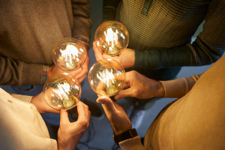 Team of four intelligent people together holding bright, shining, glowing Edison light bulbs as...