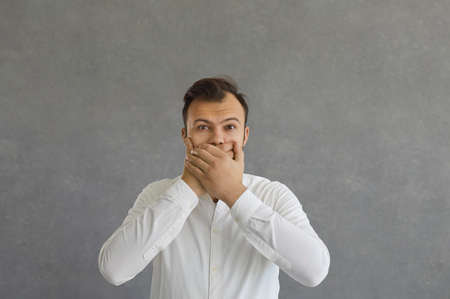 Do not say anything. Surprised and shocked young caucasian man frightenedly closes his mouth with...