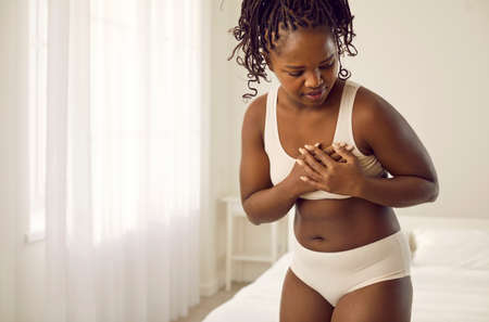 Beautiful young black woman in underwear touches chest when she suddenly feels pangs of pain in heart or breast. Copy space home background. Cardiac pain, cardiovascular disease.の写真素材
