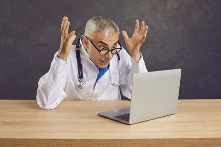 Crazy angry senior male doctor screaming in frustration looking at laptop screen. Frustrated gray-haired man in glasses and in medical gown with stethoscope sits at table on gray background.の写真素材