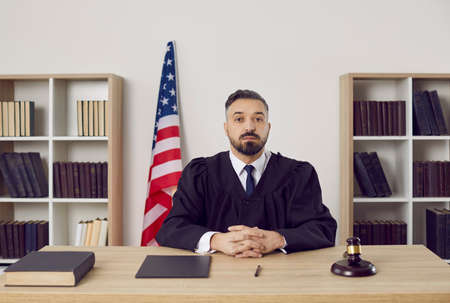 Portrait of a young American judge dressed in a robe or gown sitting at his table with books and gavel in the courtroom during a court hearing in the courthouse. Law and justice in the US conceptの写真素材