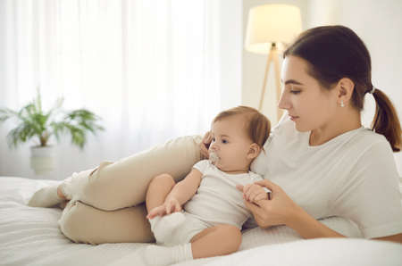 Loving young Caucasian mother relax on bed at home cuddle with baby infant. Caring happy mom rest...
