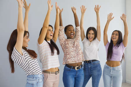 Overjoyed millennial gen z diverse multiracial girls friends raise hands up celebrate shared success. Smiling excited young women have fun feel euphoric triumph with win or victory. Luck concept.の写真素材