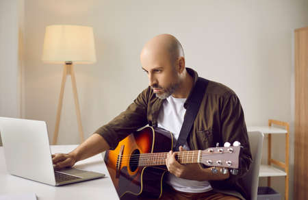 Serious focused beginner musician learning to play music on his instrument at home, looking at laptop computer screen, searching for guitar tab lessons and chords of modern rock or country compositionの写真素材