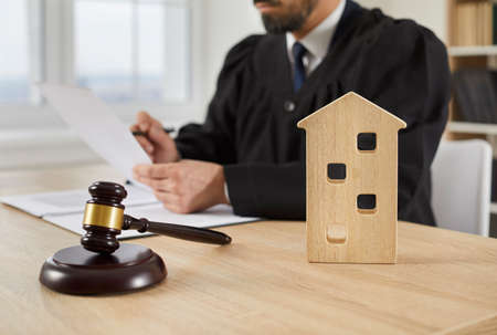 Close up of a gavel and a small symbolic toy house on a wooden table in court, with a judge working with settlement documents in the background. Real estate law, property, foreclosure conceptsの写真素材