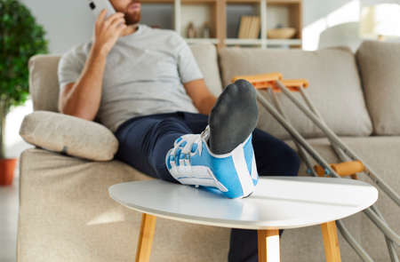 Close up of injured man with bandage on leg sit on sofa talk on cellphone with doctor. Unhealthy guy...