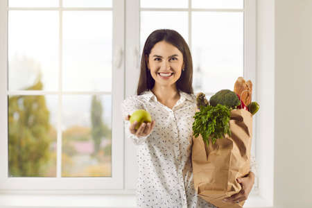 Pretty young woman with her groceries at home. Happy beautiful lady standing in the kitchen with paper grocery bag full of healthy foods, smiling, looking at camera and holding out fresh tasty appleの写真素材