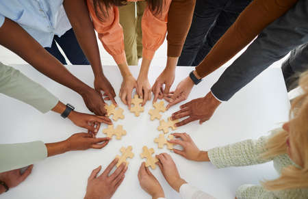 Hands of different business people connecting puzzle pieces as sign of problem solving and teamwork. Top view of multiracial people standing in circle at table with wooden puzzles. Business concept.の写真素材