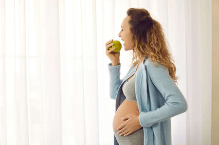 Happy beautiful young pregnant woman with big belly standing by copyspace window and enjoying fresh tasty green apple, profile view. Mothers health, eating good healthy food during pregnancy conceptの写真素材
