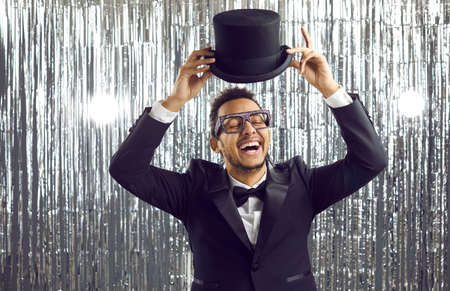 Positive funny young man wearing glasses and modern party tuxedo standing on silver foil fringe background, feeling free, excited, happy and carefree, having fun, taking off his top hat and laughingの写真素材
