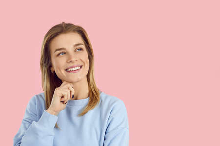 Happy dreamy woman imagines something pleasant on pastel pink text background. Dreamer takes decision. Creative confident teenage girl in blue sweatshirt thinking hand on chin of modern youth solutionの写真素材