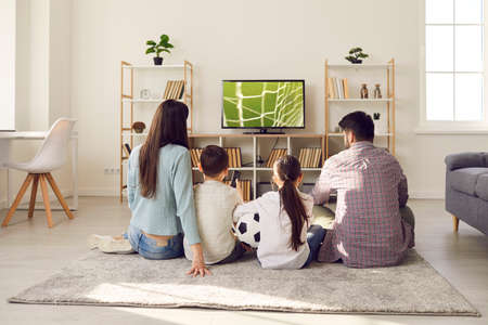 Young family with children watching football match together at home cheering for their favorite team. Mom, dad, daughter and son are sitting on floor in front of big TV with their backs to camera.の写真素材