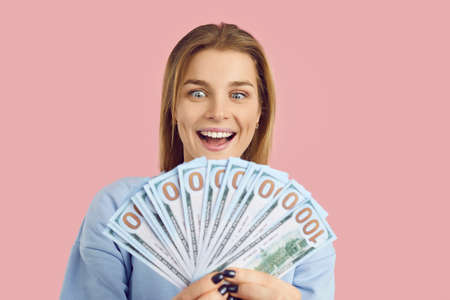 Wealthy woman isolated on pink background smiling and holding bunch of paper banknotes in her hands....