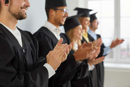 Happy, smiling college or university graduates in hats and robes standing in row and clapping hands....