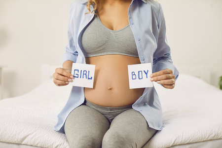 Pregnant woman holding paper cards with inscriptions boy and girl near her bare belly. Cropped image...