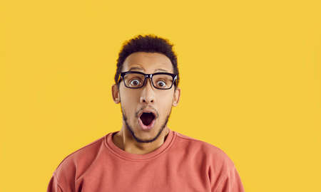Young dark skinned man with funny surprised and shocked facial expression isolated on yellow background. Close up of guy with his mouth wide open from shock of unexpected news or information.の写真素材