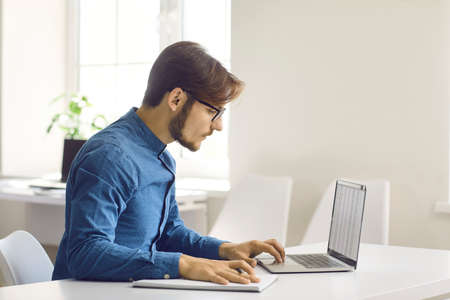 Focused millennial Caucasian man sit at desk work online on computer make notes write. Young...