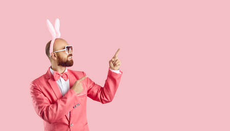 Funny young man wearing easter bunny ears shows on copy space on pastel pink background. Cheerful man in glasses and pink suit advertises or recommends. Creative banner for celebration of Easter.の写真素材