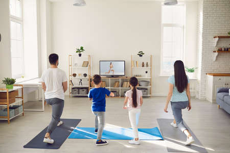 Family having fitness workout at home. Back view of happy mum, dad and kids standing on rubber mats together, watching video workout with gym instructor on TV screen and doing leg stretching exerciseの写真素材