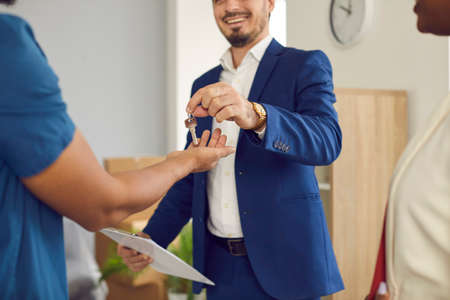 Male home salesman, real estate agent or realtor gives keys to new home to young married couple. Cropped image of man in formal suit handing over keys to new owners of apartments. Real estate concept.の写真素材