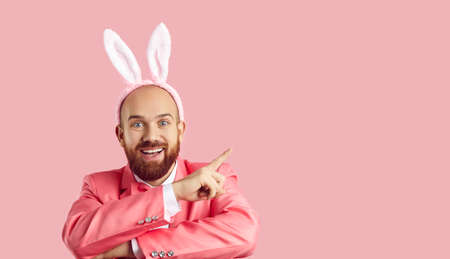 Look here, guys. Funny happy cheerful young man with ginger beard wearing pink suit and plush bunny ears pointing index finger to side at something on blank advertising copyspace Easter backgroundの写真素材
