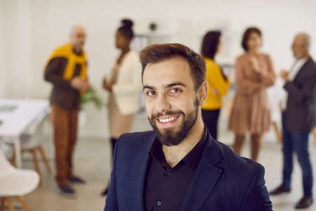 Portrait of handsome and successful businessman on office background in modern business center. Head shot of confident smiling millennial Caucasian man standing against backdrop of colleagues.の写真素材