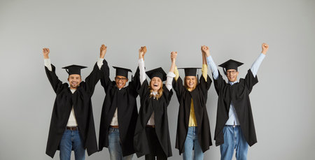 Portrait of happy and successful group of multiracial graduates standing in row holding hands. People in mantles and mortarboards on gray background. Concept of education, graduation and people.の写真素材
