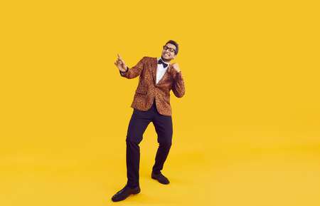 Happy young black man in party outfit dancing isolated on yellow colour background. Full length shot of positive guy in leopard jacket, bowtie and glasses enjoying music and having fun in the studioの写真素材