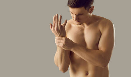 Muscular athletic man experiencing sharp pain in his wrists due to sports injury isolated on gray background. Serious naked man holding his hand near copy space. Medical banner.の写真素材