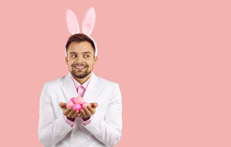 Happy cheerful handsome young man in white suit and cute plush bunny ears standing on pink background, holding handful of dyed Easter eggs, smiling and looking away at blank copyspace on right sideの写真素材