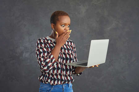 Emotional dark skinned woman with puzzled and shocked expression looks at laptop in her hands. Young woman stands on a gray background and sees the shocking content on the screen.の写真素材