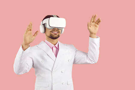 Happy smiling man in suit and VR gear headset experiencing virtual reality and exploring AR cyberspace environment of new videogame. Studio shot on pink color background. Innovation, future conceptの写真素材