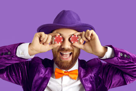 Studio portrait of happy goofy excited man with ginger beard, in purple suit, hat and orange bow tie smiling and covering both his eyes with two red poker chips in hands. Casino, gambling, fun conceptの写真素材