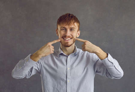 Portrait of young man wearing casual shirt showing and pointing with fingers to white teeth and...