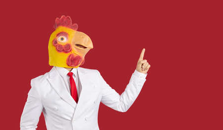 Crazy man wearing white suit and funny chicken rooster mask standing isolated on solid dark red colour background and pointing index finger at blank marketing copy space on right sideの写真素材