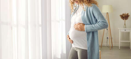Young happy pregnant woman in comfortable home clothes standing by bright window at home holding her hands on her tummy. Pregnancy, maternity preparation and expectation concept. Cropped image. Bannerの写真素材