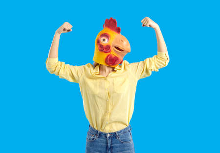 Woman in funny chicken disguise pretends to be super strong. Portrait of confident young girl wearing strange weird ridiculous absurd bizarre Hallowen mask flexing her arms isolated on blue backgroundの写真素材