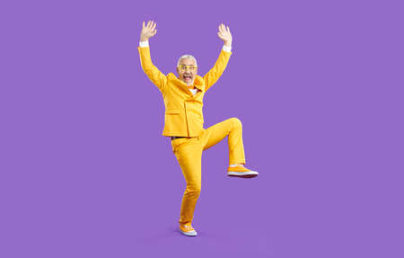 Funny old man having fun and fooling around in the studio. Happy crazy senior man wearing a bright yellow party suit and glasses standing with his hands raised up isolated on a solid purple backgroundの写真素材