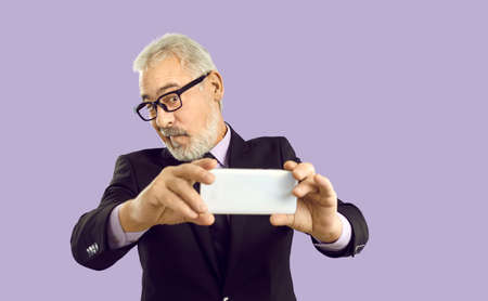 Senior businessman making funny face and taking selfie on mobile phone. Studio shot of handsome...