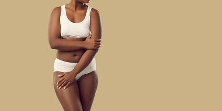 Unrecognizable dark-skinned young woman with fuller body type in lingerie isolated on beige background. Plus size female model in white set of lingerie slightly covers body standing near copy space.の写真素材