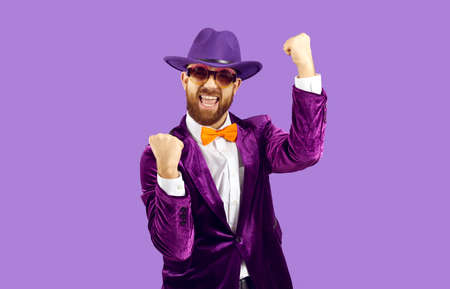 Happy confident man celebrating success. Funny excited cheerful bearded guy in hat, suit and orange bow tie standing on purple background, fist pumping and shouting Yes, Yay, I did it, Cool, Hurrayの写真素材