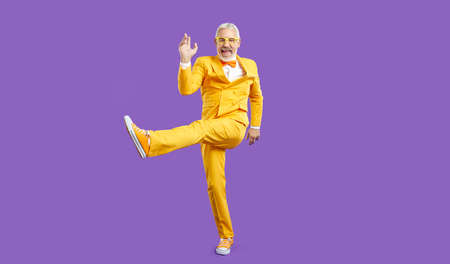Happy funny energetic stylish senior man having fun in studio. Cheerful mature guy wearing bright yellow party suit with bow tie enjoying music and dancing isolated on solid purple backgroundの写真素材