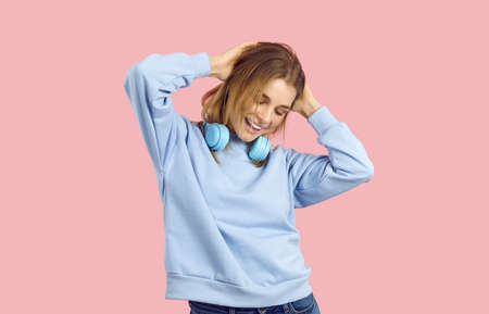 Young woman enjoying good vibes and carefree lifestyle. Pretty girl wearing trendy blue sweat shirt posing isolated on solid pastel pink, listening to cool music, dancing, touching hair, feeling happyの写真素材