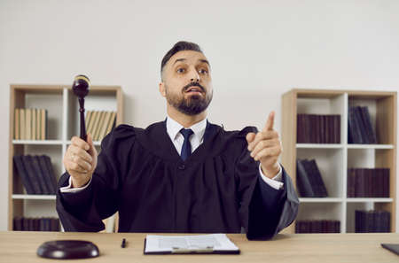 Judge in court of law considers the case closed and passes judgement. Young man in a robe uniform sitting at a table in the courtroom pronounces a fair sentence and hits the sound block with a gavelの写真素材