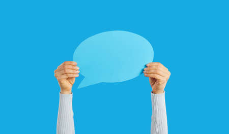 Unrecognizable woman holding empty blue speech bubble in hand on blue background. Blank paper card layout with copy space for message, quote, inspiration, opinion or vote. For advertisementの写真素材
