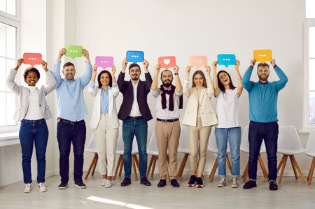 Happy young people holding colorful paper message bubbles. Diverse group of cheerful male and female workers standing, showing mobile messenger chat icons and sharing dialogues, replies and reactionsの写真素材