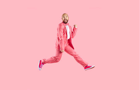 Happy excited funny young man in suit and sneakers having fun jumping on pink background. Cheerful Caucasian bald bearded man in pink formal suit is having fun and jumping. Banner. Full length.の写真素材