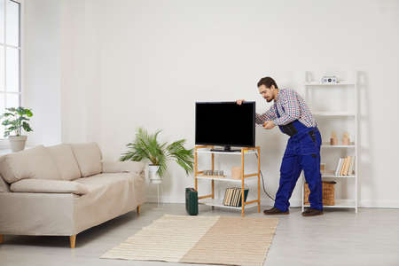 Young professional male TV technician installs and connects new TV in customers house. Repairman in overalls uses screwdriver to screw frame to TV screen. Concept of calling repairman at home.の写真素材